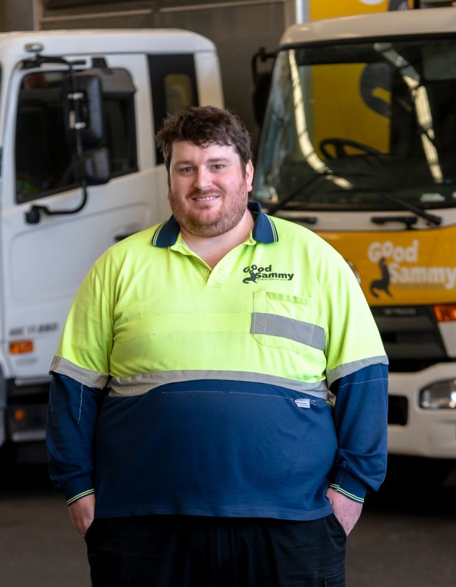 Sam wearing Good Sammy hi-vis uniform, standing in front of trucks with hands in pocket and smiling at camera.