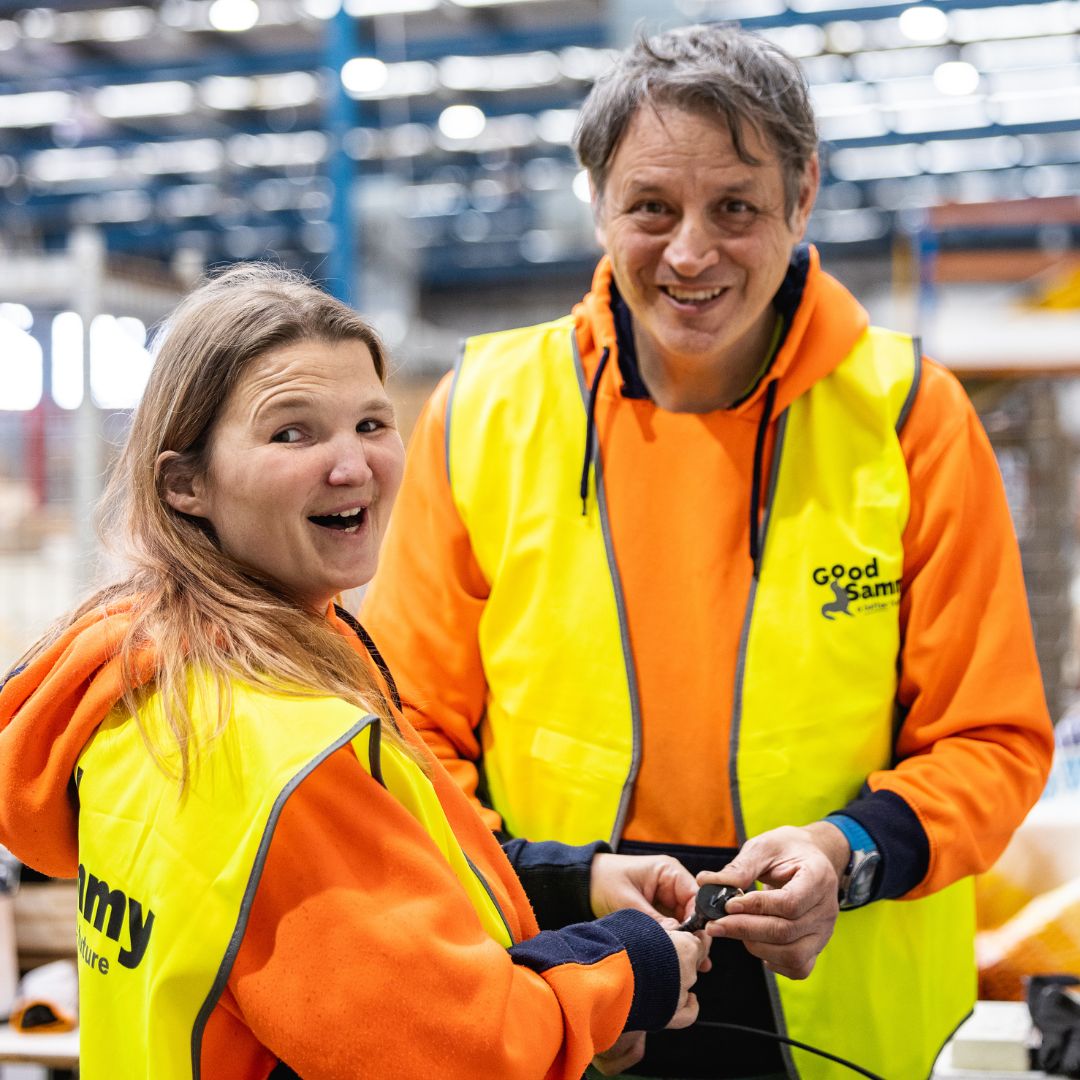 Image of two Good Sammy staff members wearing bright orange hoodies and yellow high vis vests sorting electrical equipment.