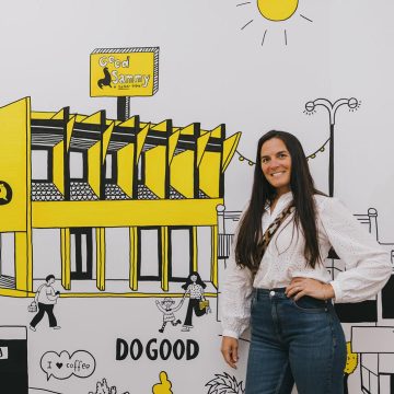 A woman standing in front of a mural depicting the Good Sammy Leederville store in bright yellow, with black accents.
