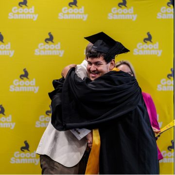 A person wearing a black graduation gown and cap, and yellow sash, is hugging another person in celebration.