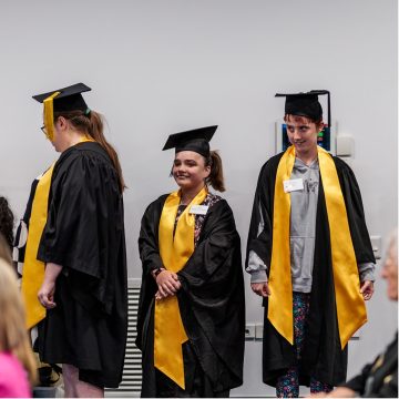 Three people standing in a row, looking away from the camera. All three are wearing black graduation gowns and caps, and yellow sashes.