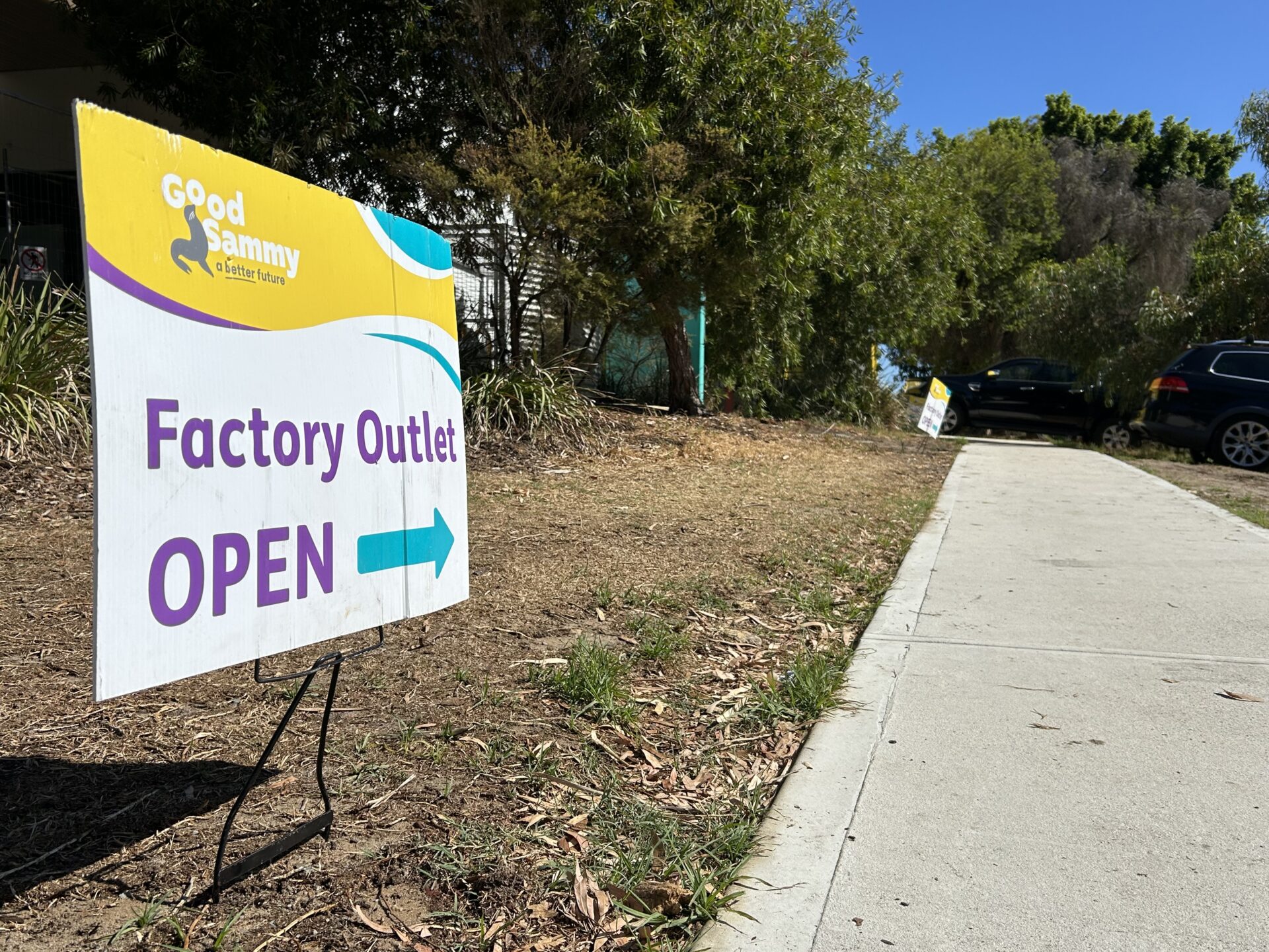 A sign that says 'Good Sammy Factory Outlet Open', staked into the grass, next to a footpath that leads into the distance.