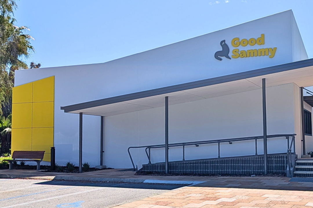Photo of outside of newly constructed Good Sammy building. The building is white with a yellow Good Sammy logo at the top. The entrance to the building includes both stairs and a ramp.